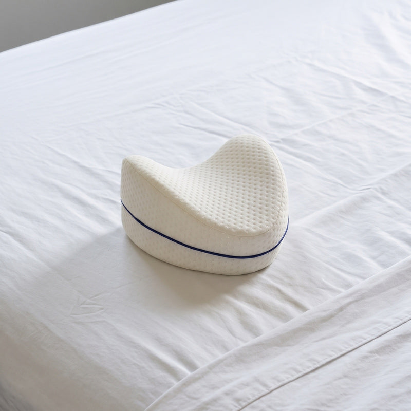 Cojín Alinea sobre una cama con sábanas blancas: forma de gota maciza, cara superior cóncava, funda blanca de jacquard con vivo azul marino.
